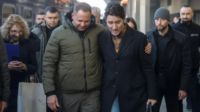 Ермак в зимней одежде приобнимает Трюда в пальто за плечо. За ними несколько человек, одетых для холодной погоды. На фоне виден поезд.