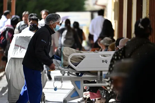 Un enfermero con tapabocas y malla de pelo lleva a un paciente en una camilla por un corredor de un hospital en el que hay mucha gente