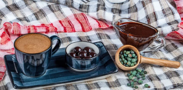 Na kraciastym obrusie stoi mała ciemnoniebieska taca, a na niej filiżanka i miseczka do kompletu. Filiżanka jest pełna kawy, a miseczka ciemnych owoców przypominających czarne oliwki. Na prawo widać małą drewnianą chochlę, a w niej zielono-niebieskie owoce menengiç. Część z nich wysypała się na obrus. Nieco dalej stoi szklany dzbanek zawierający ciemnobrązowy sos.