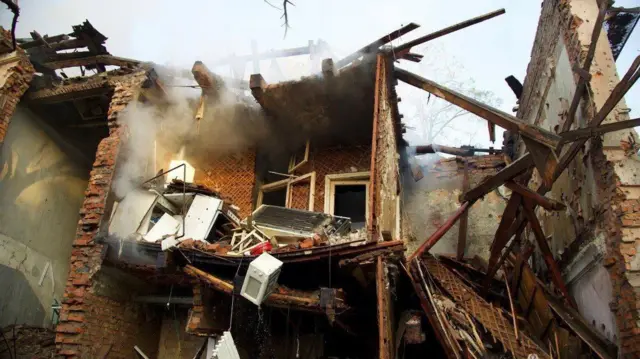 Зруйнований приватний будинок: обвалені стіни й дах, видно оголені цегляні конструкції та уламки меблів. З другого поверху звисає мікрохвильовка. Усередині та навколо — багато завалів, із пошкодженого приміщення піднімається дим.