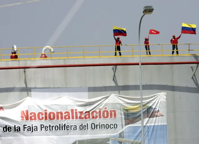 Trabajadores petroleros ondean banderas de Venezuela en la cima de un tanque de petróleo en el que hay una gran pancarta que celebra la "nacionalización de la Faja Petrolífera del Orinoco".