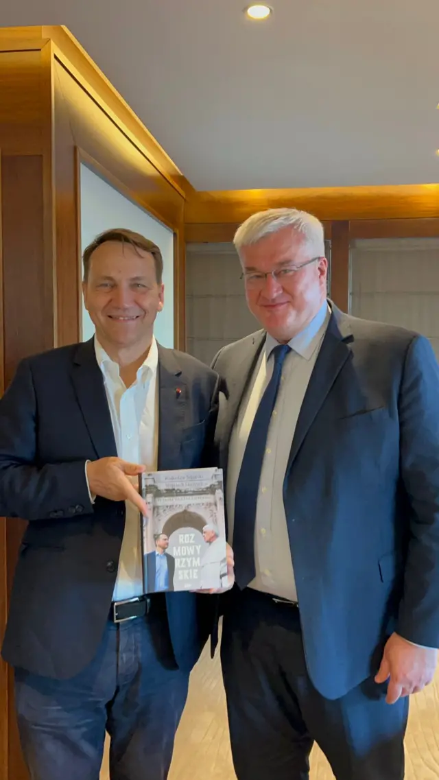 Сікорський і Сибіга стоять поруч у приміщенні та тримають книгу, показуючи її обкладинку. На обкладинці видно назву "Rozmowy Rzymskie", а позаду – дерев’яні стіни й освітлення стелі.