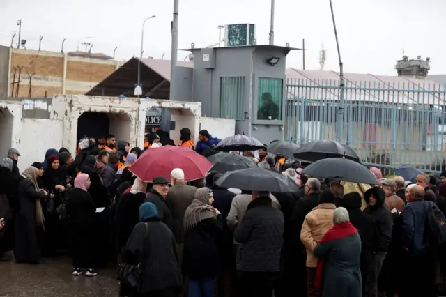 جمع مكتظ لنساء ورجال فلسطينيين قرب حاجز قلنديا العسكري، وعدد منهم يحمل مظليات. الأجواء ماطرة.