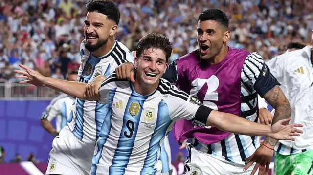 Julian Alvarez celebrates a goal with arms stretched out and a big smile as Argentina team-mates join in behind him. Alvarez is wearing Argentina's blue and white striped shirt.