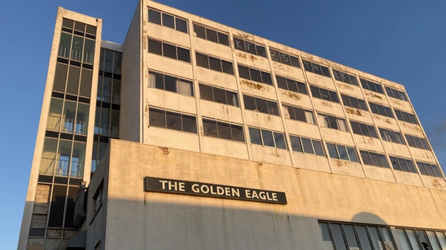 El exterior del hotel de hormigón. Hay un gran letrero que dice "El Águila Dorada". Es un edificio de varias plantas. A un lado está la escalera. Parece muy ruinoso.  