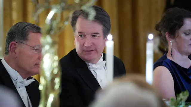 Фрагмент урочистої вечері: люди у фраках і вечірніх сукнях сидять за довгим столом, прикрашеним високими свічками та квітами. Інтер’єр у золотистих тонах.