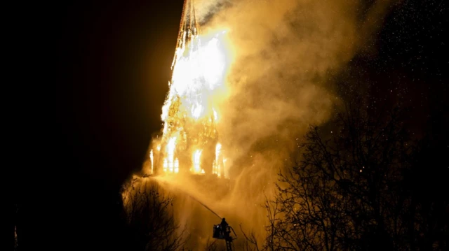Kilisenin kulesi alevler içinde, bir itfaiye eri gece karanlığında su sıkıyor.