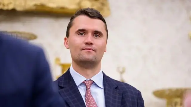 Charlie Kirk em pé, com sorriso contido, dentro de salão