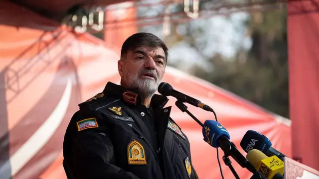 24 Kasım 2025'te, açık hava sahnesinde, kırmızı bir arka plan önünde duran Tuğgeneral Ahmed-Rıza Radan, bir grup mikrofona konuşuyor.