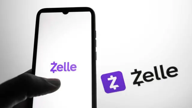 Uma mão segurando um celular com o logotipo do aplicativo Zelle na tela, com letras roxas sobre um fundo branco. Ao fundo, é possível ver o mesmo logotipo, nas cores preto e roxo