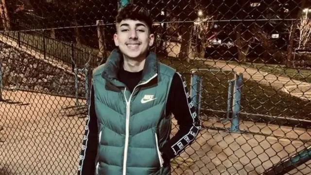 Batın Barlasçeki, bir parktaki spor aletlerinin etrafına çekilmiş tellerin önünde, üzerinde siyah bir bluz ve yeşil bir yelekle kameraya bakarak gülümserken