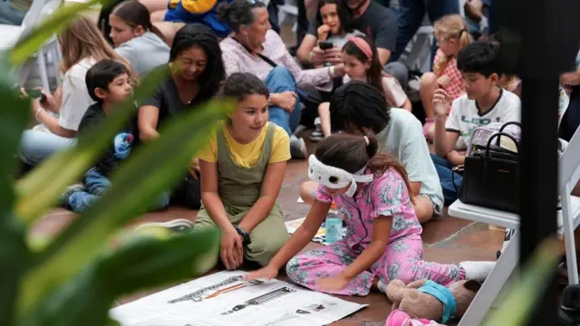 Dve devojčice sede na podu okružene drugom decom i gledaju u crtež makete rakete i letelice Orion
