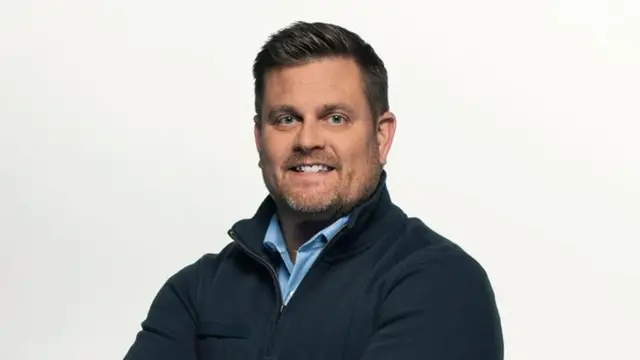 A foto mostra um homem, Brian Thompson, sorrindo, de braços cruzados, usando uma camisa azul-clara sob uma jaqueta escura, diante de um fundo branco.