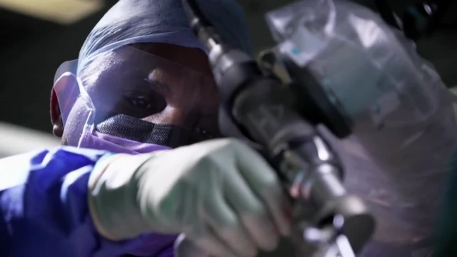 Se ve a Sivakumar Shankar con una mascarilla quirúrgica, gorra y guantes mientras mira a través de un equipo médico mientras lo sostiene con una mano.