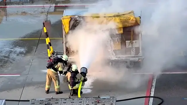 На фото – двоє пожежників розпилюють піну на вантажний візок в аеропорту. З нього піднімається дим