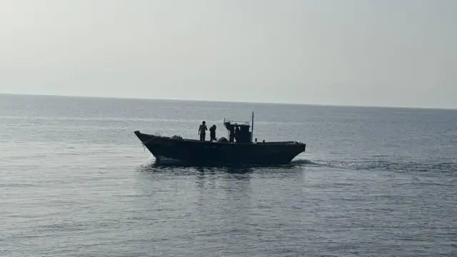 木造船が海に浮かんでいる。船の上に2人が立っているのが見える