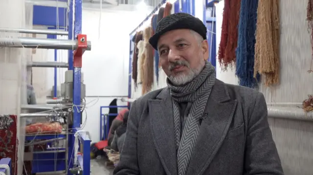 نثار احمد حسینی، مدیر عامل شرکت قالین‌بافی «المک بافت» با بالاپوش خاکستری و دستمال خاکستری و کلاه سیاه در یک کارگاه قالین بافی.