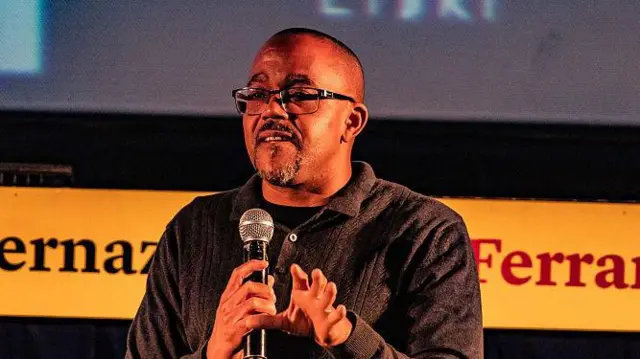 Kehinde Andrews, one black man wey wear dark shirt, square-framed glasses and one black and grey beard, hold microphone.