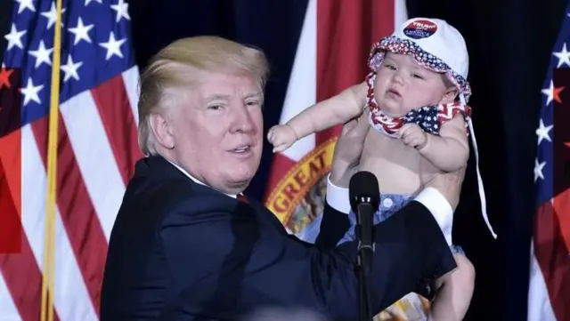 ترامپ در یک مراسم، پشت تریبون، نوزادی را رو به جمعیت بالا گرفته است
