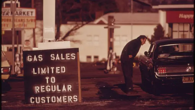 Imagem de um cartaz no lado externo de um posto de gasolina nos Estados Unidos, durante a crise do petróleo de 1973, dizendo 'Venda de gasolina limitada a clientes regulares'. Ao fundo, pode-se ver um homem inclinado em direção à janela de um carro, conversando com o motorista.