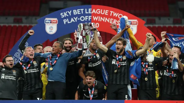 Un grupo de personas con camisetas de fútbol a rayas negras sostiene un pequeño trofeo con aire feliz. Al fondo, una pancarta roja y azul con la leyenda "Ganador del Play-Off de la Sky Bet League Two 2020".