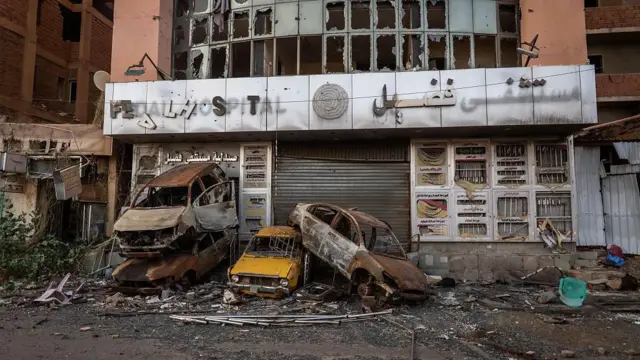 Des véhicules calcinés sont empilés devant un bâtiment abandonné, détruit par le conflit. Les fenêtres sont soufflées et un panneau brisé indique « hospital » en anglais.