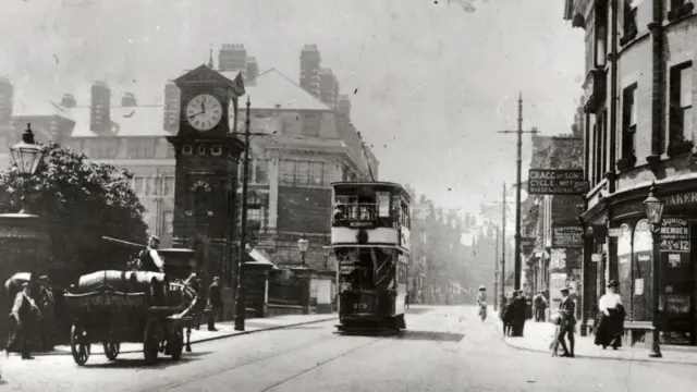 Fotografía de época en blanco y negro de Stamford New Road en Altrincham, procedente del archivo del Ayuntamiento de Trafford. A la izquierda se ve una torre de reloj, un carro tirado por un caballo a un lado de la calle mientras un tranvía circula por el centro. A la derecha, un hombre de traje espera para cruzar la calle, y una mujer con falda hasta los tobillos y camisa blanca camina por la acera.