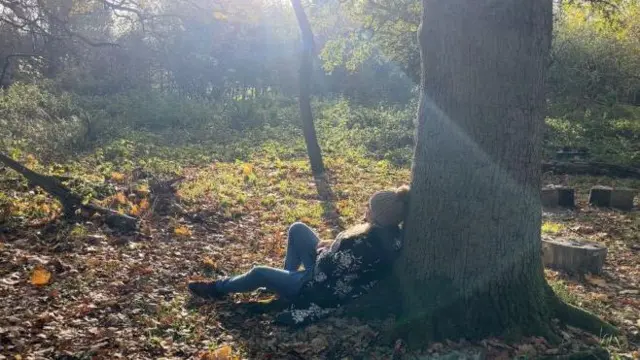 Una mujer estaba sentada con la espalda apoyada contra un árbol mirando a la distancia.