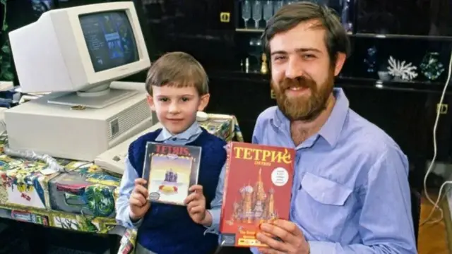 Alexey e o filho pequeno sorrindo e mostrando caixabahia esportes da sorteTetris, pertobahia esportes da sortecomputadorAlexey Pajitnov and a boy holding Tetris boxes. in front of a 1980s computer