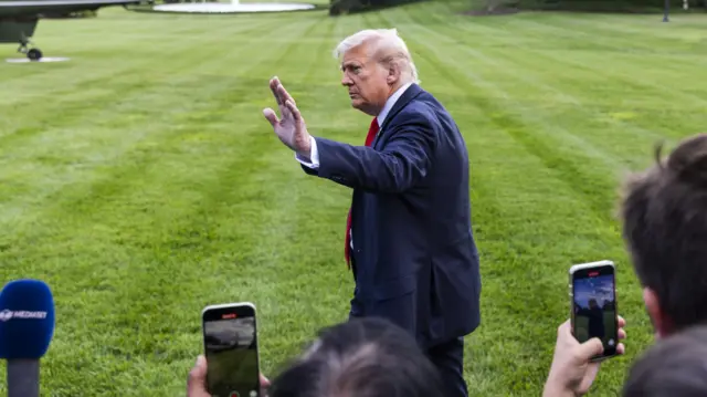 Дональд Трамп на фоне зеленого поля в синем пиджаке и красном галстуке стоит вполоборота и машет журналистам рукой. Его снимают на телефоны