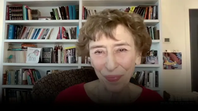 Женщина сидит в своем кабинете на фоне книжных полок, она улыбается