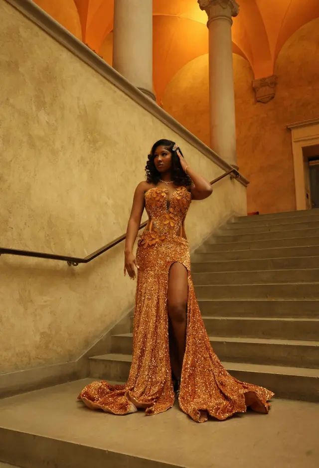 Anita Konneh porte une robe ocre à paillettes avec une fente au niveau de la jambe. Elle se tient debout sur un escalier - des colonnes gréco-romaines peuvent être vues derrière elle.