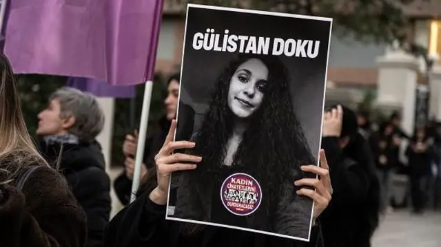 Gülistan Doku'nun posterini tutan bir kadın eli merkezde, geride kadınlar bir yürüyüşte görünüyor, mor bir bayrak var, flular.