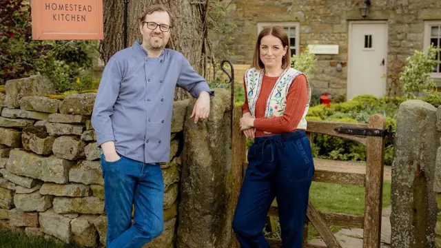 Peter y Cecily se encuentran afuera de una cabaña de piedra tradicional, cerca de una puerta de madera colocada en un muro de piedra seca.