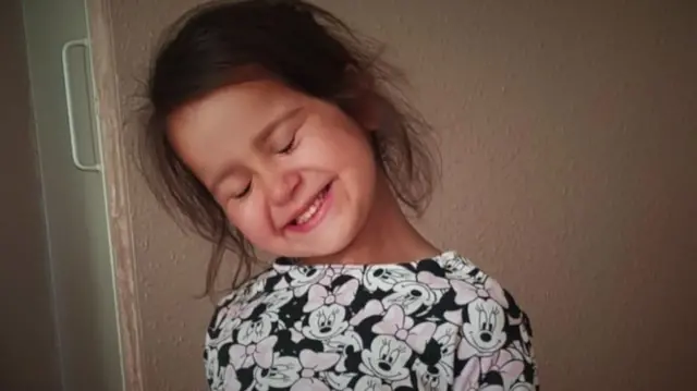 Sara Sharif, uma menina de pele clara e cabelos castanhos com uma camiseta da minnie, sorrindo de olhos fechados 