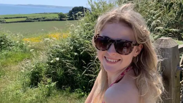 Picture of Eve Jones, as she stands by a wooden gate overlooking rolling green hills in front of the sea. She wears a pink halterneck top and looks over her shoulder smiling with her teeth. She has light blonde mid length hair, she wears square tortoise shell sunglasses with silver small hoops. 