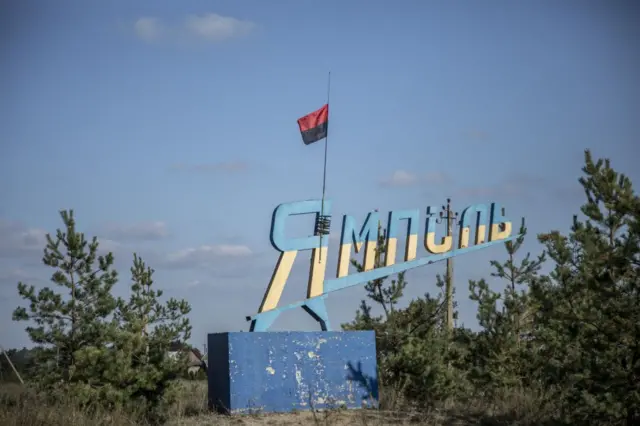 Надпись «Ямполь» на въезде в населенный пункт. Покрашено в сине-желтый, к ней прицеплен красно-черный флаг.