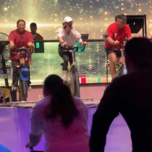Three people ride exercise bikes on a stage, illuminated by bright lights. We see the silhouettes of two guests who are riding bikes and facing the instructors. 