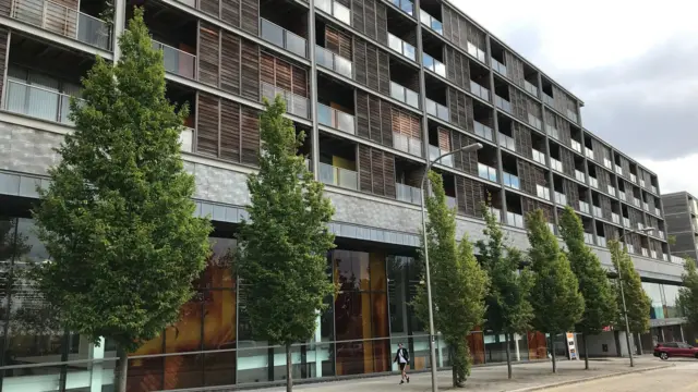 Una imagen de una parte del complejo Vizion. Es un gran bloque de pisos con lo que parece ser un revestimiento de madera alrededor de los balcones. La planta baja alberga una gran tienda. Frente al edificio hay una hilera de árboles y una acera. Se puede ver a una persona caminando por el sendero.