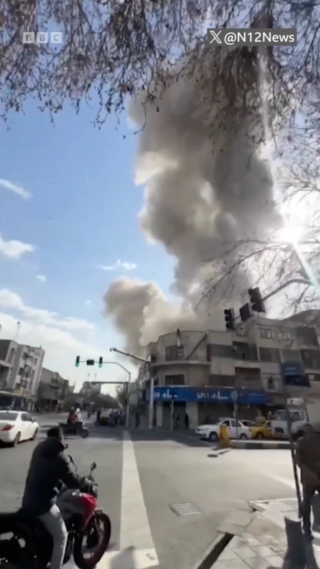 Міське перехрестя, над яким здіймається великий стовп світлого густого диму. Будівлі вздовж вулиці виглядають пошкодженими або в зоні недавнього вибуху. На передньому плані – дорожній рух: мотоцикліст, кілька автомобілів і перехожі, які зупинилися та дивляться у бік диму.