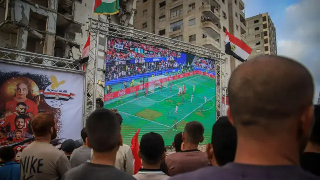 تجمع فلسطينيون يحملون الأعلام لمشاهدة مباراة المجموعة السابعة من الدور الأول لكأس العالم لكرة القدم 2026 بين مصر وإيران على شاشة عملاقة نُصبت بين أنقاض المباني المدمرة، وذلك على الرغم من استمرار الهجمات الإسرائيلية على مدينة غزة في 27 يونيو/حزيران 2026.