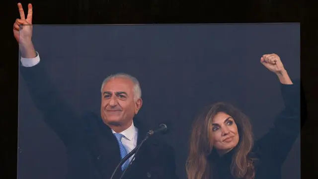 رضا پهلوی، ولیعهد پیشین ایران و از چهرههای شاخص اپوزیسیون کنونی، در حالی که در کنار همسرش یاسمین پهلوی روی صحنه ایستاده، در جریان تجمع مخالفان ایرانی در ۱۴ فوریه ۲۰۲۶ در محل نمایشگاه ترزینویزه در مونیخِ جنوب آلمان، همزمان با برگزاری شصتودومین کنفرانس امنیتی مونیخ، علامت پیروزی نشان میدهد.