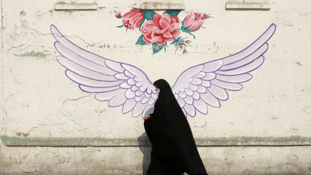 زنی با چادر از جلوی دیواری که بر آن بال فرشته نقاشی شده رد می‌شود