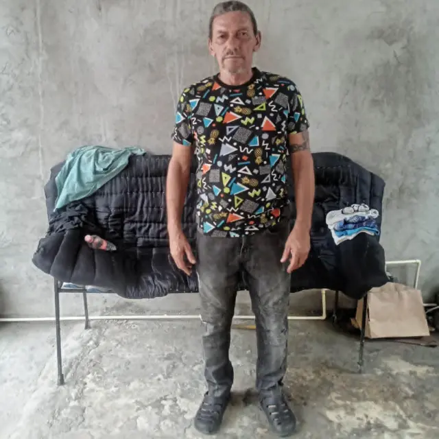 Lázaro Romero León, de pé, olha para a câmera em frente a um pequeno sofá, com jeans escuro e uma camiseta estampada, em um pátio de cimento na cidade de Tapachula, no Estado mexicano de Chiapas