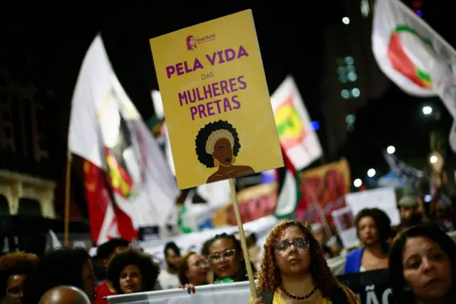 Mulheres negras protestando à noite; uma delas levanta cartaz que diz 'Pela vida das mulheres pretas'