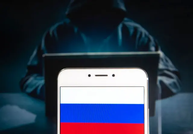 Стоковое фото: некто в капюшоне в темной комнате за компьютером, на переднем плане смартфон с российским флагом