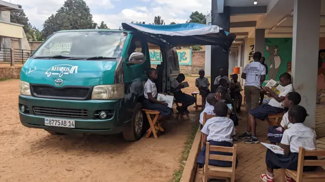Des élèves assis à côté du bus transformé en bibliothèque mobile stationnée dans la cour de l’école primaire d’application ISP Bukavu.