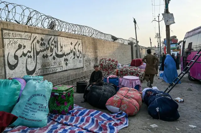 کودک افغان نشسته روی اثاثیه مهاجران در کنار اتوبوس‌ها