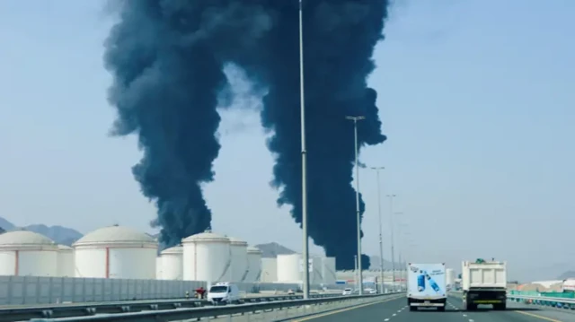یک اتوبان که دو ستون عظیم دود از تاسیسات نفتی برخاسته، چند خودرو در حال عبور و مرور