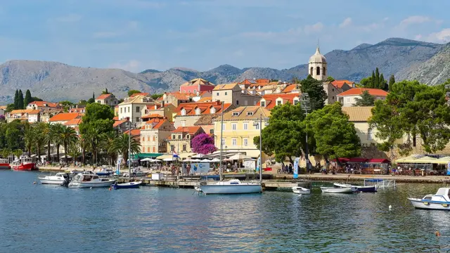 Мальовниче фото прибережного міста з води, будинки з жовтими та червоними дахами, зелені дерева, човни біля пристані, на задньому плані гори